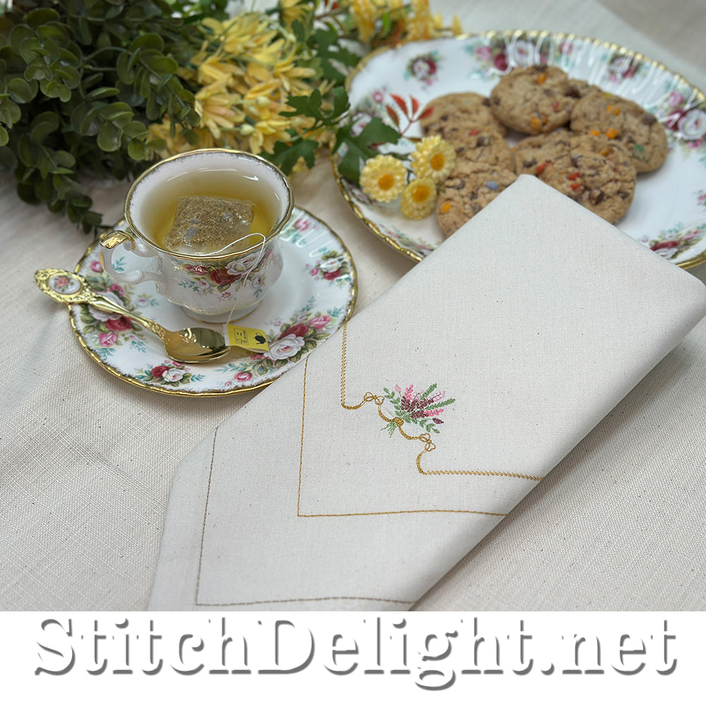 Teacup with floral saucer, embroidered napkin, and cookies on a table with 'StitchDelight.net' branding.