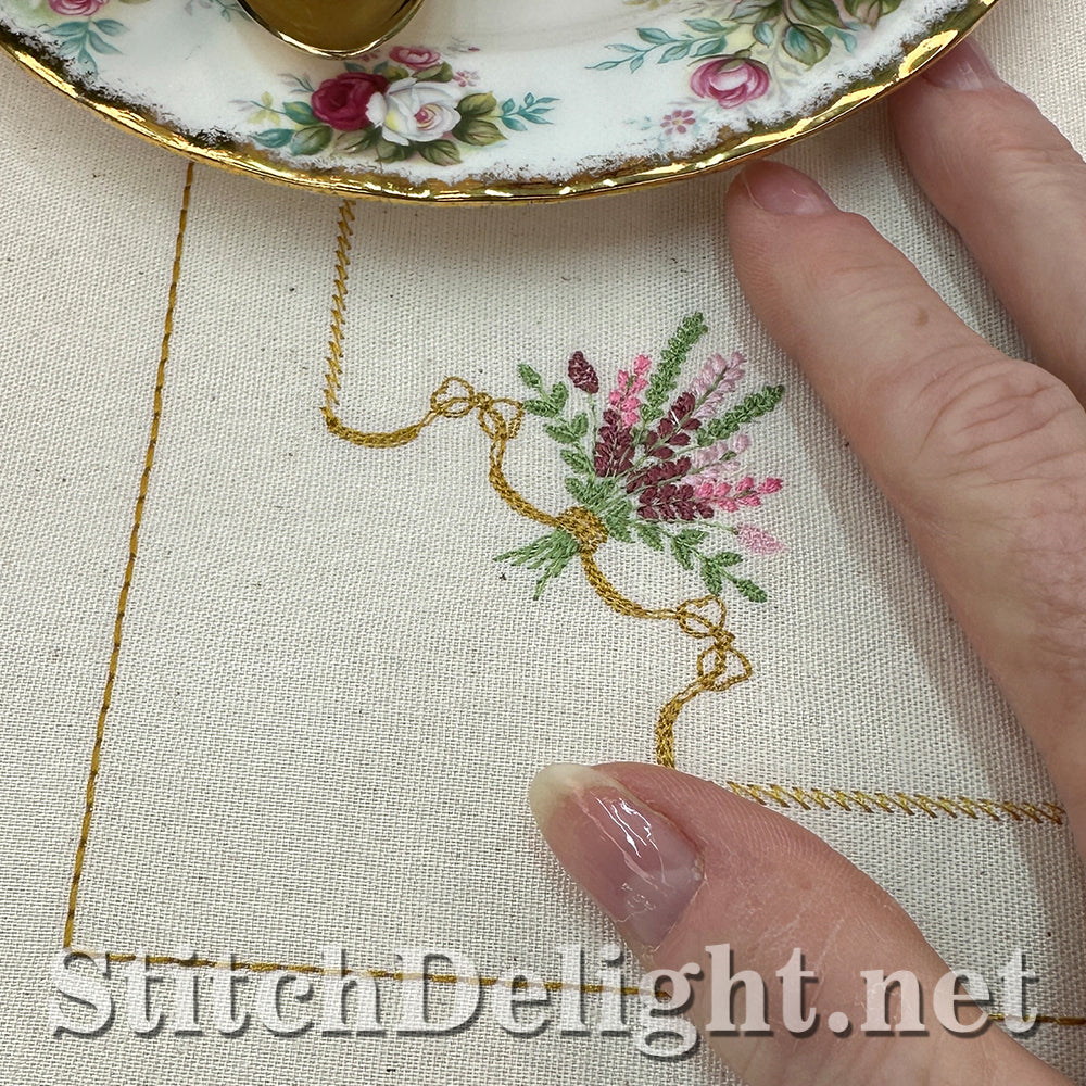 Close-up of embroidery with floral design and hand holding a teacup, brand 'StitchDelight.net' visible.