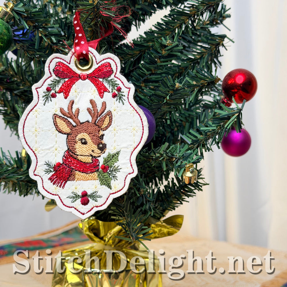 Close-up of the embroidered deer tag with antlers, red scarf, holly and berries, a red bow and a gold eyelet hanging on a miniature Christmas tree