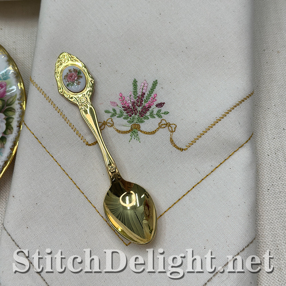 Gold spoon with decorative handle on a piece of fabric with floral embroidery, featuring 'StitchDelight.net' branding.