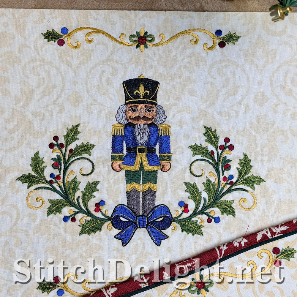 The blue colorway: a nutcracker in a blue coat with a blue bow between holly sprays, with the gold scroll border above