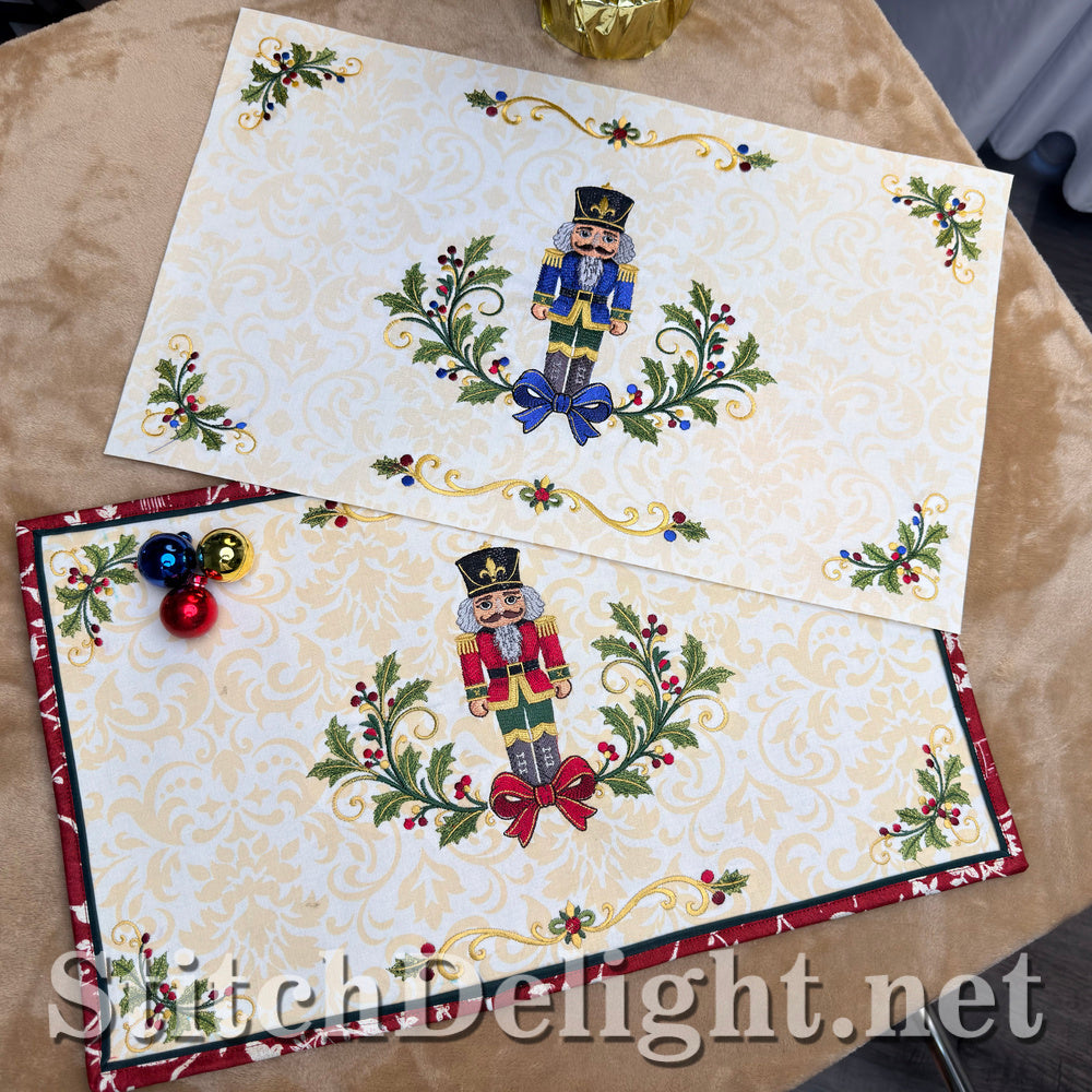 Two finished Christmas placemats in the red and blue colorways, each with a nutcracker between holly sprays, scroll borders, holly corners and a red flange binding